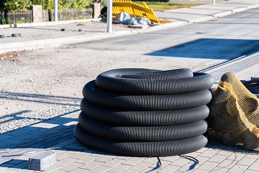 Black corrugated pipe rolled into a coil at a construction site. Freshly paved sidewalk and road construction.