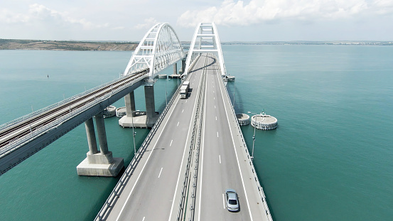 Aerial view along the long beautiful bridge above turquoise sea. Action. Flying above bending bridge with driving cars
