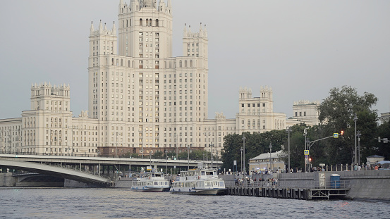 One of Seven Sisters, beautiful skyscraper in the Stalinist style, Moscow river and touristic ships. Action. Concept of architecture, city embankment with walking people