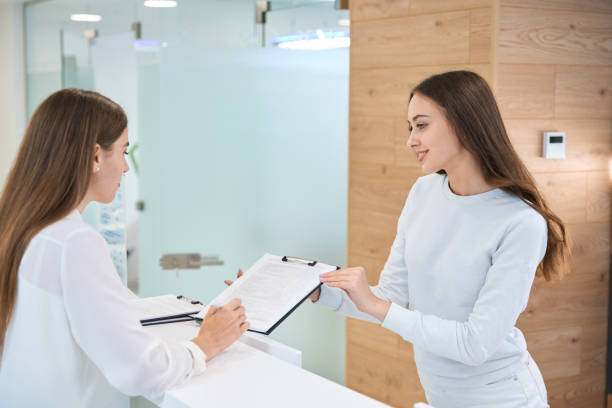 istock Female patient signing document at front desk assisted by secretary 1445836480
