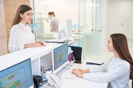 Young client standing at reception counter and looking at clinic receptionist seated at desktop computer