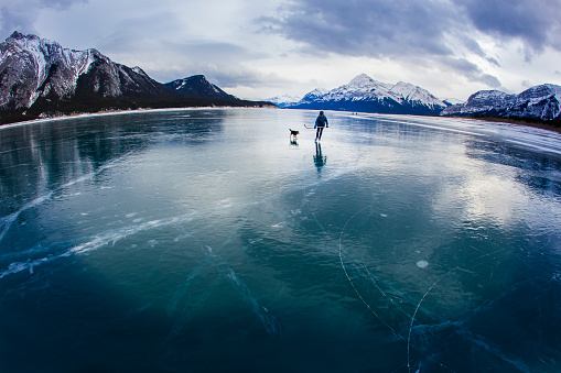 A woman goes for an ice skate on a frozen lake with her dog in the Rocky Mountains of Canada. She carries a hockey stick and wears a hockey helmet.