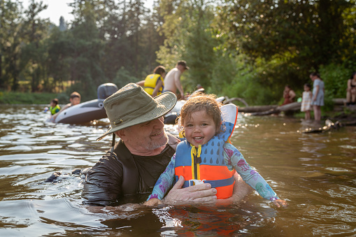 A cute multiracial toddler girl wearing a lifejacket and swimsuit laughs while swimming with her grandfather in the river on a warm and sunny afternoon. The multi generation family is on summer vacation.