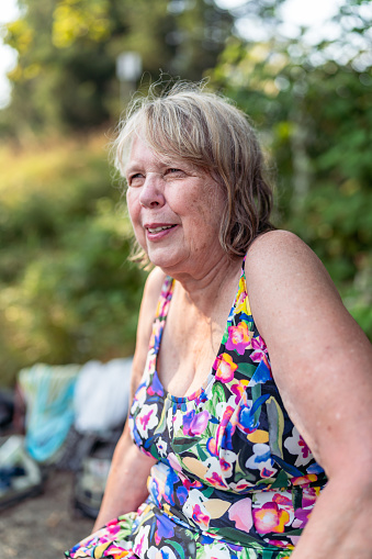 A mature adult woman wearing a colorful floral swimsuit sits outdoors at the river while on a relaxing summer vacation with her family.