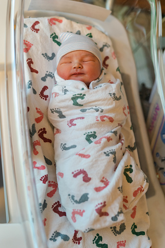 High angle view looking down at an adorable Eurasian newborn baby boy wearing a knit hat and swaddled in a receiving blanket with a colorful footprint pattern peacefully sleeping in a bassinet at the hospital.
