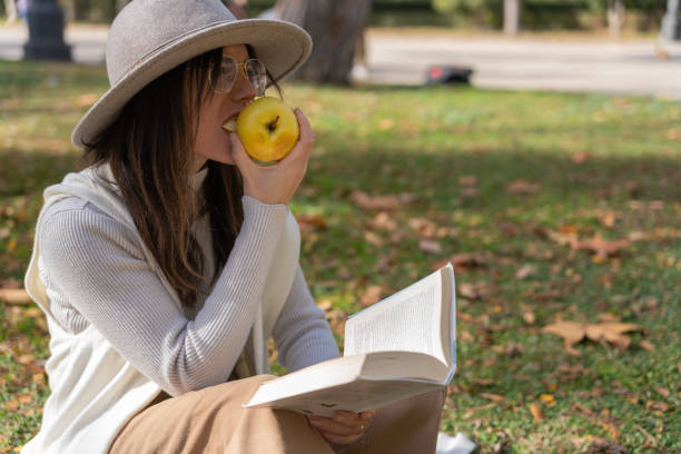 une femme brune au chapeau mange une pomme verte tout en tenant un livre ouvert avec ses mains - book brown apple education photos et images de collection