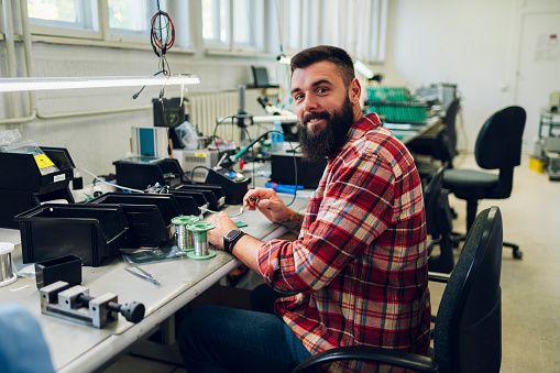 Portrait of an electronics technician is soldering circuit board in his workshop and looking into the camera. Computer support engineer. Modern technology. Repairing by hand. Wearing checkered shirt.