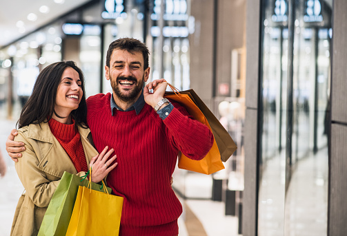 Young attractive happy couple hugging, smiling and holding shopping bags while walking in shopping mall