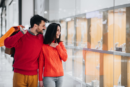 Lovely young couple of newlyweds on a shopping spree in a mall, looking at a store window. Handsome young man hugging his lovely girlfriend in a mall.