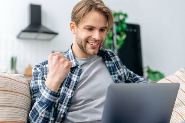 Close-up excited caucasian guy, freelancer, sits on a sofa in living room, looks at laptop screen in amazement, gesticulates with fist, celebrates big profit, received good news, smiling, yes gesture primer plano emocionado chico caucásico, profesional independiente, se sienta en un sofá en la sala de estar, mira la pantalla de la computadora portátil con asombro, gesticula con el puño, celebra grandes ganancias, recibió buenas noticias, sonriendo