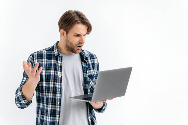 istock Frustrated confused puzzled caucasian stylish young man, standing on a white isolated background, holds an open laptop, surprised looks at the screen, see unexpected news, message, gesturing with hand 1445855932