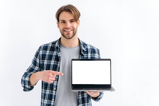 istock Stylish handsome confident caucasian man, standing on a white isolated background, holds an open laptop, showing empty white mock-up screen for advertising or presentation, points finger at it, smile 1445855941