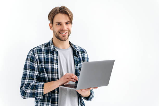 istock Positive handsome confident caucasian stylish young man, freelancer, student, programmer, standing on a white isolated background, holds an open laptop, looking to the camera, smiling friendly 1445855943