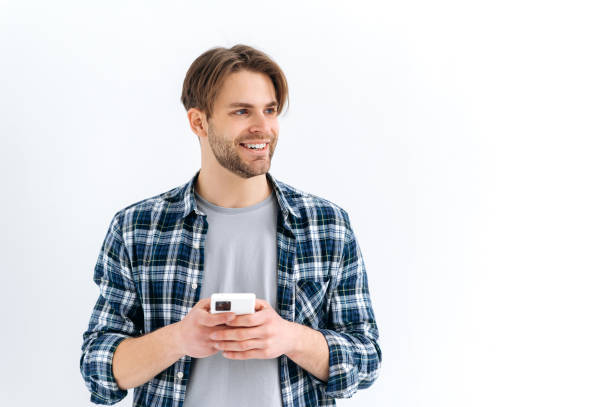 istock Positive handsome stylish caucasian guy, holding smartphone in hand, chatting online, browsing internet, looking to the side, thinking, dreaming, standing on isolated white background, smiling 1445855953