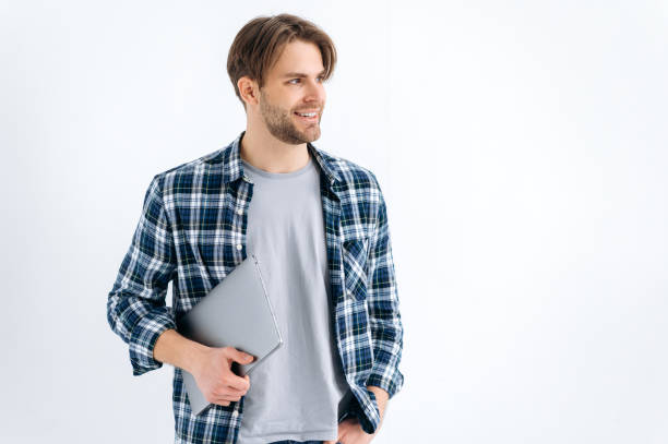 istock Trendy handsome confident caucasian stylish young man, freelancer, student, programmer, standing on a white isolated background, holds laptop, looking to the side, smiling friendly. Copy-space 1445855958