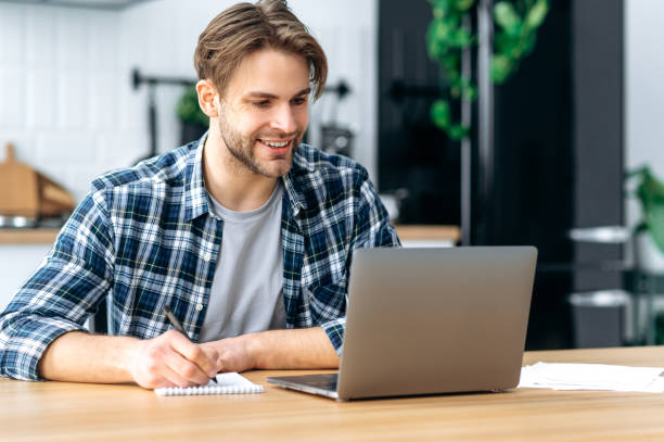 istock Modern clever caucasian young man, student, freelancer, creative manager, sitting at home, taking notes in a notebook during an online webinar or lecture, working on a project, using a laptop, smiling 1445855993