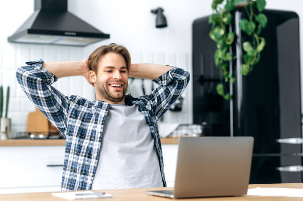 istock Calm satisfied caucasian guy, freelancer, working from home, sits in the kitchen at workplace, takes a break from work, puts his hands behind his head, looks to the screen, glad with a project, smiles 1445856012