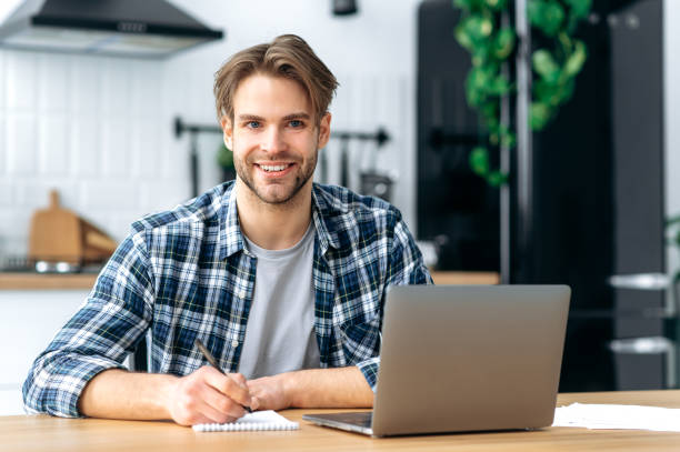 istock Successful clever caucasian young man, student, freelancer, programmer, sitting at home in kitchen, taking notes in a notebook during an online webinar or lecture, working on a project, using laptop 1445856014