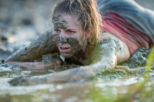 A young woman is seen crawling through a large mud puddle as she competes in a Mud Run.  She is dressed comfortably in athletic wear and has a focused expression on her face.