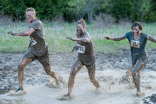 A small team of three competitors are seen running through a large puddle of water as they take part in a Mud Run.  They are each dressed comfortably in athletic wear and have exhausted expressions on their faces.