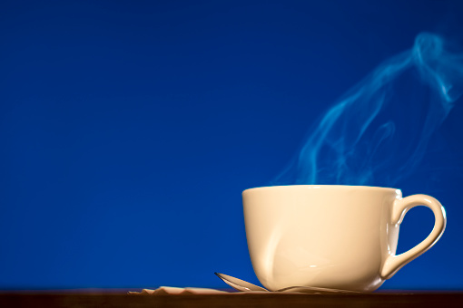 A low angle view of steam rising from a large white porcelain cup of hot coffee or tea sitting with a silver spoon on a wooden table against a blue background.