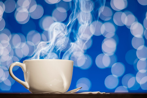 Steam rising from a large white porcelain cup of hot coffee or tea sitting with a silver spoon on a wooden table against a blue background with white bokeh.