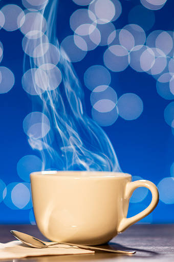 A low angle view of steam rising from a large white porcelain cup of hot coffee or tea sitting with a silver spoon on a wooden table against a blue background with white bokeh.