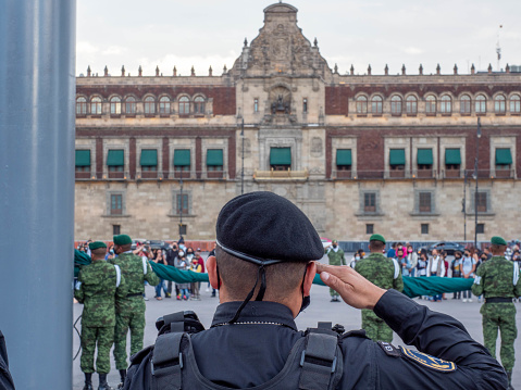 Mexico City, Mexico - February 11,  2022: A Mexican Police officer maintains salute as soldiers wrap the Mexican flag during the Zocalo Square Flag Ceremony