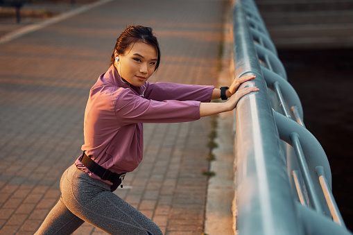 Young Asian athletic woman warming up during sports training outdoors and looking at camera.