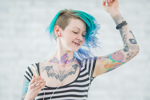 A young hip woman with body piercings and tattoos, poses for a portrait.  She is dressed casually in a striped t-shirt and has her hair dyed and Ombre blue and green as she listens to music on her cell phone and dances around.