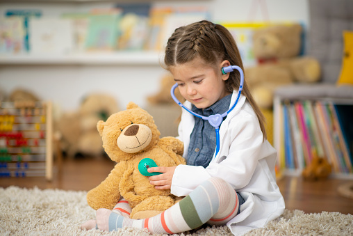 A sweet little girl sits on the floor at daycare as she plays dress up with a doctors lab coat and a stethoscope.  She is holding the stethoscope to the teddy bears chest as she pretends to listen to his heart.