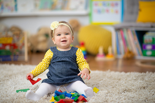 A sweet little baby sits on the floor of her daycare classroom as she plays with colorful wooden blocks.  She is dressed casually and smiling as she plays joyfully.