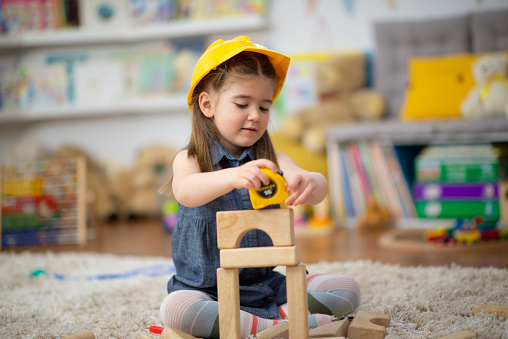 A sweet little girl sits on the floor at daycare as she dresses up in a hardhat and uses a measuring tape to measure her block structure.  She is dressed casually and is focused n her pretend game of construction.