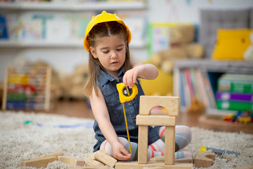 A sweet little girl sits on the floor at daycare as she dresses up in a hardhat and uses a measuring tape to measure her block structure.  She is dressed casually and is focused n her pretend game of construction.