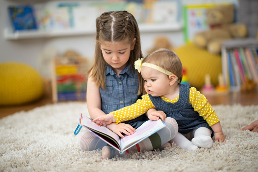 Two sweet little sisters sit on the floor together at daycare with a book open in front of them.  The older sister is readying to her baby sister as they enjoy the story.  They are both dressed casually and are focused on the pictures.