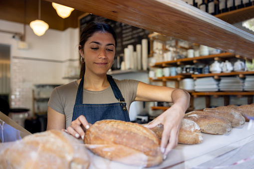 Latin American woman working at a bakery and placing bread loafs on the shelf - small business concepts