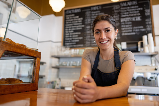 Happy Latin American waitress working at a cafe and leaning on the bar counter while looking at the camera