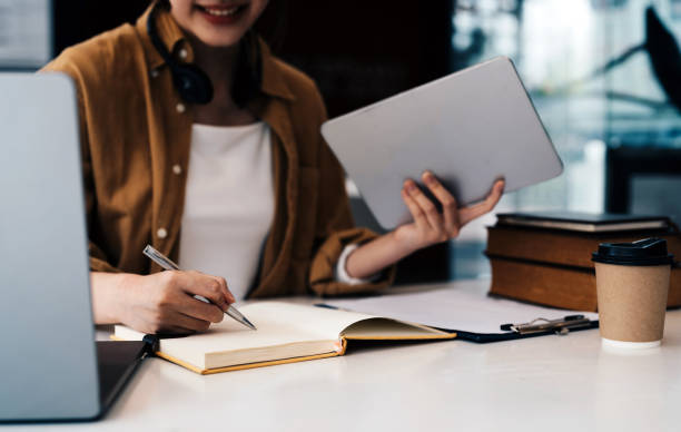 istock Young asian woman using laptop and writing making list taking notes in notepad working or learning on laptop indoors- educational course or training, seminar, education online concept. 1445899434