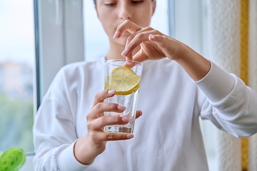 Close-up of glass of water with lemon in hands of young female. Healthy drink, refreshing vitamin beverage
