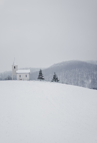 A vertical shot of a small white church on top of a snowy hill in Northern Bosnia and Herzegovina.