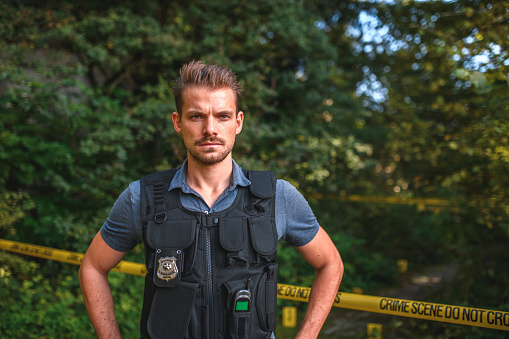 Portrait of a serious Caucasian detective in protective vest standing arms akimbo and looking at the camera, barricade tape and evidence tags in the background with trees. Waist up image, looking at the camera.
