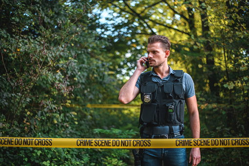 Armed Caucasian crime investigator in protective wear, talking on the phone, between  yellow police tapes. Wide angle shot, 3/4  length image.