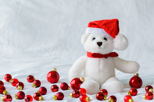Selective focus on Santa claus teddy bear eyes who wearing hat sitting with red Christmas baubles on white cloth background.
