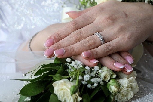 A closeup shot of bride hands with pink nail polish and a sparkly ring over a white green bouquet