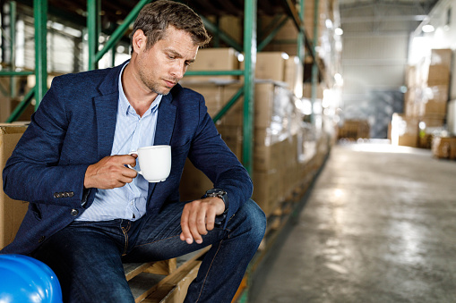 Mid adult foreman checking the time on his wristwatch while having a coffee break in storage room. Copy space.