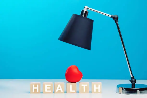 Photo of Red heart on top of wooden cubes with the word HEALTH on a table next to a black table lamp.