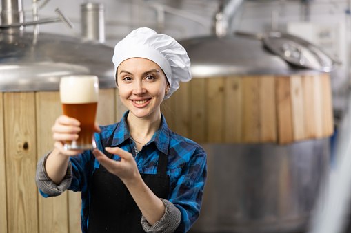 Cheerful young female brewer recommending craft beer, holding out glass with drink while standing against background of fermenters in brewery
