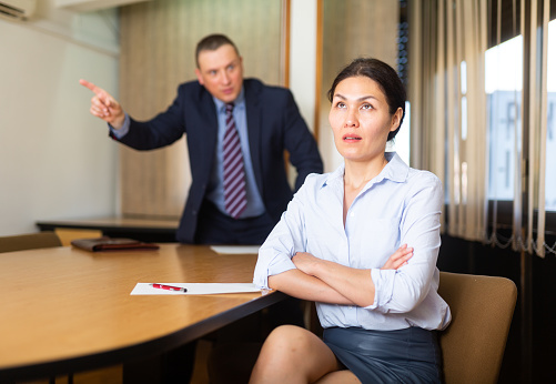 Frustrated asian female entrepreneur sitting at office desk on background with angry partner expressing his dissatisfaction, pointing out mistakes in work