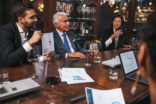 Business meeting in a luxury restaurant. Group of multi ethnic and diverse age range people attending to a corporate briefing sitting at a table after lunch.