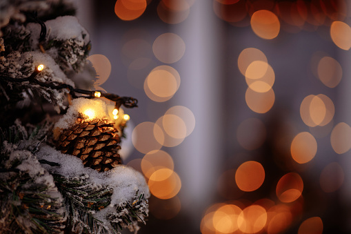 Close-up of a snow-covered Christmas tree decorated with golden balls and luminous garlands against the backdrop of a private house. Soft focus blurs.